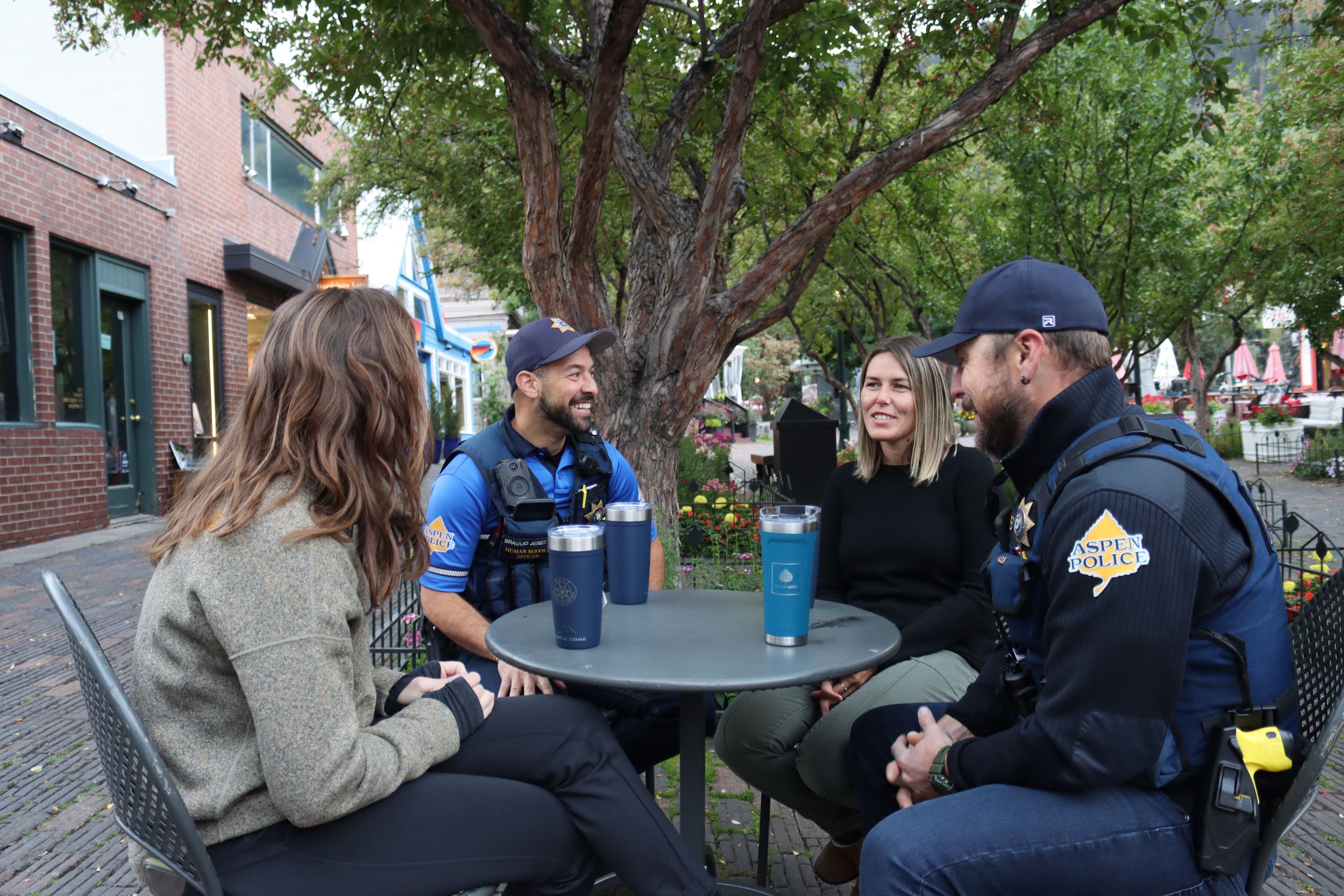 Two male police officers in blue uniforms sitting at a table talking to two women
